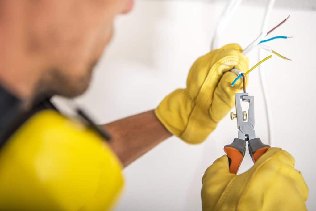 An electrician wearing yellow protective gloves uses wire-stripping pliers to remove insulation from electrical wires.