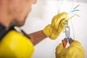 An electrician wearing yellow protective gloves uses wire-stripping pliers to remove insulation from electrical wires.