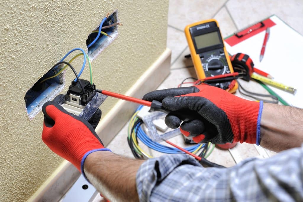 An electrician wearing protective gloves uses a screwdriver to repair an electrical outlet with exposed wiring in a wall, while a digital multimeter, measuring tape, and tools are visible nearby.