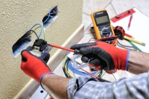An electrician wearing protective gloves uses a screwdriver to repair an electrical outlet with exposed wiring in a wall, while a digital multimeter, measuring tape, and tools are visible nearby.